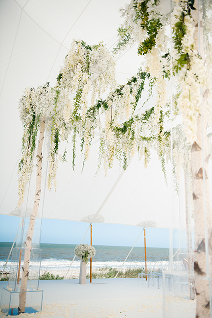 A pretty beach wedding ceremony decorated with birch poles, white flowers, and ghost chairs 