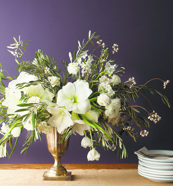 a not too Christmasy floral arrangement of silver urn with a base of olive branches and tallow berries, white amaryllis, ranunculus, and paperwhites from the Wall Street Journal