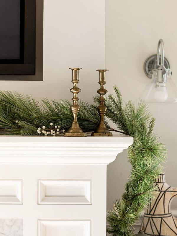mixed artificial pine garland on a mantel decorated with white berries behind two gold candlesticks as a classic christmas decor idea